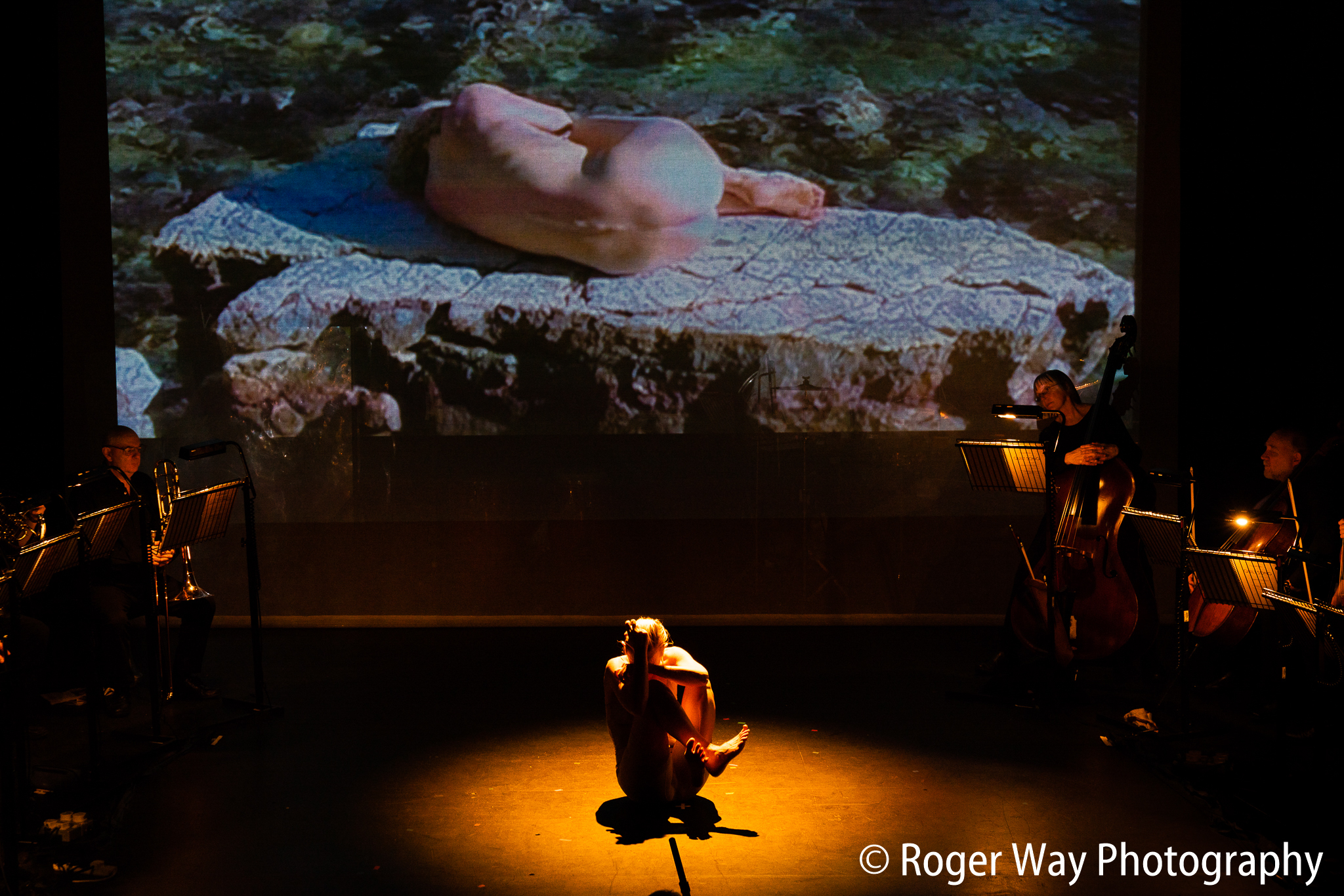 A naked performer sits in a foetal position on an empty stage.